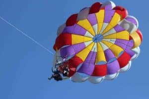 Two people parasailing beneath a multicoloured canopy against a blue sky.